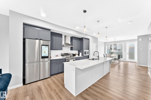 Kitchen featuring wood-finish flooring, gray cabinetry, stainless steel appliances, a white subway tile backsplash, and a large island with a sink and stone countertop - 479 39 St Sw, Edmonton, AB 