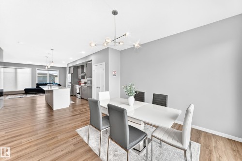 Open-concept dining and kitchen area featuring a white dining table with upholstered chairs, wood-finish flooring throughout, stainless steel appliances, white cabinetry, pendant lighting, and abundant natural light from large windows - 479 39 St Sw, Edmonton, AB 
