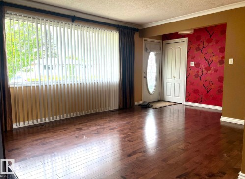 Rich wood-finish flooring throughout, featuring a wide window with vertical blinds, a front entry door with an oval glass insert, and a wall with decorative floral wallpaper - 15003 81 Street Nw, Edmonton, AB 