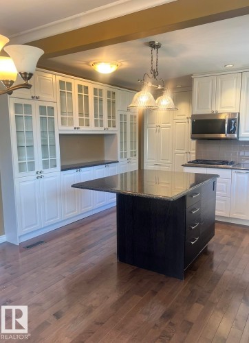 Wood-finish flooring throughout, white cabinetry with glass inserts, central island with dark finish and stone countertop, stainless steel microwave, and integrated ceiling beam - 15003 81 Street Nw, Edmonton, AB 