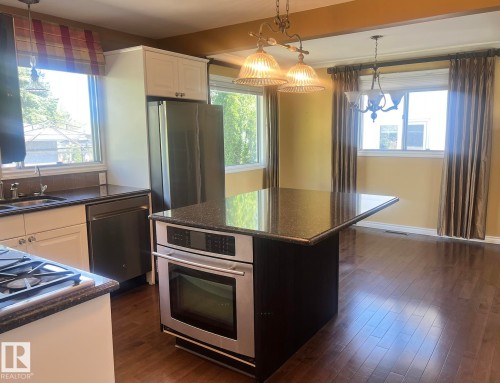Kitchen featuring wood-finish flooring, stainless steel appliances, dark countertops, white cabinetry, and an island with a built-in oven - 15003 81 Street Nw, Edmonton, AB 