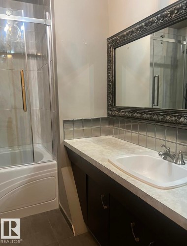 Bathtub with sliding glass enclosure, dark wood-finish vanity with an integrated sink, textured countertop, and a decorative framed mirror - 15003 81 Street Nw, Edmonton, AB 