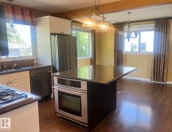 Kitchen featuring wood-finish flooring, stainless steel appliances, dark countertops, white cabinetry, and an island with a built-in oven - 