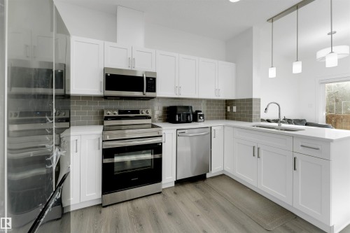 Modern kitchen featuring white cabinetry, stainless steel appliances, light-toned countertops, a subway tile backsplash, and wood-finish flooring - #62 2710 66 St Sw, Edmonton, AB 