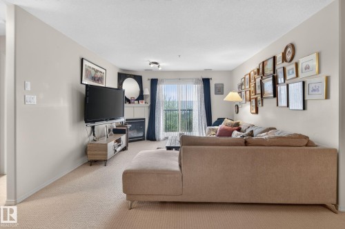 Living room featuring light-colored wall-to-wall carpeting, a built-in fireplace with white mantelpiece, a sliding glass door with sheer and dark blue curtains, and track lighting - #335 2096 Blackmud Creek Dr Sw, Edmonton, AB - Indoor Photo Showing Living Room