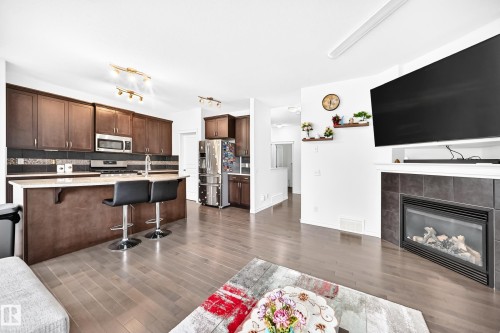 Open-concept living space with wood-finish flooring, a kitchen island with seating, dark wood cabinetry, a stainless steel refrigerator, and a fireplace with a tiled surround - 2015 Chalmers Way Sw, Edmonton, AB 