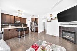 Open-concept living space with wood-finish flooring, a kitchen island with seating, dark wood cabinetry, a stainless steel refrigerator, and a fireplace with a tiled surround - 