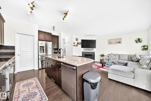 Open-concept living area featuring wood-finish flooring, a kitchen island with stone countertop, stainless steel appliances, dark wood cabinetry, and a corner fireplace - 2015 Chalmers Way Sw, Edmonton, AB 