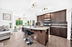 Open-concept living area featuring wood-finish flooring, a kitchen island with stone countertop, stainless steel appliances, dark wood-finish cabinetry, and multiple windows - 