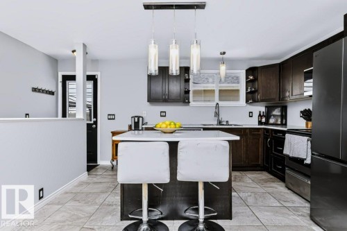 Kitchen featuring dark wood-finish cabinetry, light-toned tile flooring, a central island with a solid surface countertop, and pendant lighting fixtures - 18431 95A Avenue Nw, Edmonton, AB 