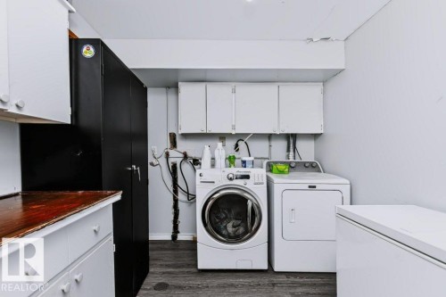 Dedicated laundry area featuring a front-load washer, dryer, wood-finish flooring, white upper cabinetry, and a built-in dark storage cabinet - 18431 95A Avenue Nw, Edmonton, AB 