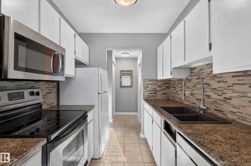 Galley kitchen featuring white cabinetry, stainless steel appliances, a mosaic tile backsplash, and a double basin sink with an extendable faucet - #204 7407 171 St Nw, Edmonton, AB 