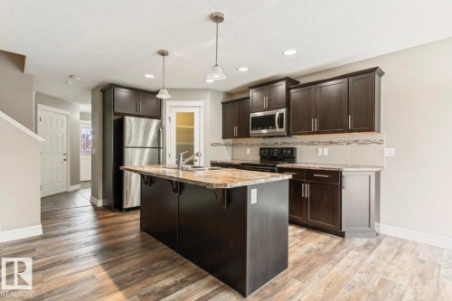 Contemporary kitchen featuring dark wood-finish cabinetry, a large center island with a stone-finish countertop, and stainless steel appliances - 79 Catalina Crest, Fort Saskatchewan, AB - Indoor Photo Showing Kitchen With Upgraded Kitchen