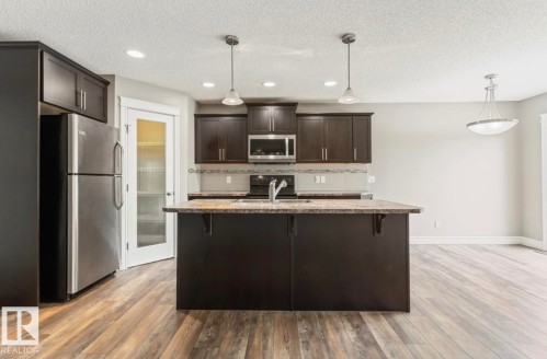 Open-concept kitchen featuring an island with integrated sink, dark wood-finish cabinetry, stainless steel appliances, and wood-finish flooring - 79 Catalina Crest, Fort Saskatchewan, AB - Indoor Photo Showing Kitchen With Upgraded Kitchen