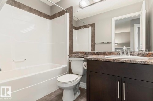 Bathroom featuring a white tub and shower surround with dark accent tiling, a dark wood-finish vanity with a light-toned countertop and integrated sink, a chrome faucet, and a large wall-mounted mirror - 79 Catalina Crest, Fort Saskatchewan, AB - Indoor Photo Showing Bathroom