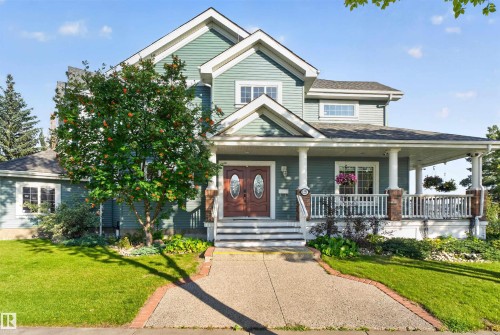 Wood-paneled double entry doors, covered porch with white columns, brick accents on porch pillars, upper-level dormer window, and a concrete pathway with brick edging - 1251 Summerside Drive Sw, Edmonton, AB 