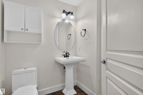 Pedestal sink with black-finish faucet, oval mirror, wall-mounted light fixture, white wall cabinet, and dark-finish flooring - 1251 Summerside Drive Sw, Edmonton, AB 