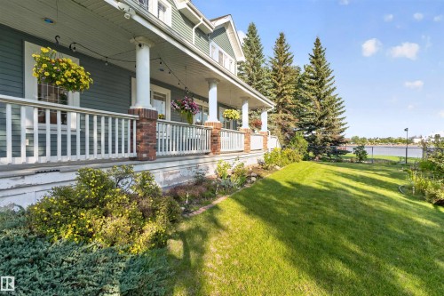 Expansive lawn area with mature evergreen trees, a well-defined garden bed, and a covered porch featuring white railings and brick-wrapped columns - 1251 Summerside Drive Sw, Edmonton, AB 
