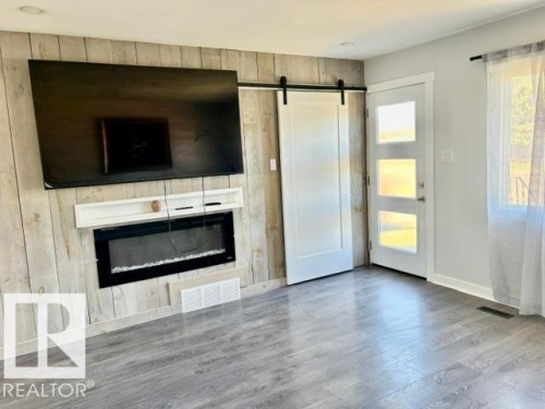 Wood-finish flooring, accent wall with a mounted electric fireplace, white barn door, and a multi-paneled entry door - 5614 50 Street, Vegreville, AB 