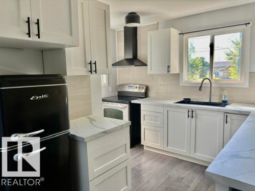 White cabinetry with matte black hardware, light-toned wood-finish flooring, light-toned backsplash, light-toned countertops, and a stainless steel sink with a dark faucet - 5614 50 Street, Vegreville, AB 