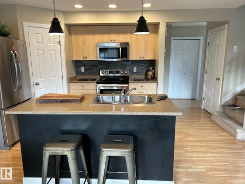 Kitchen featuring a central island with a double basin sink, stainless steel appliances, light wood-finish cabinetry, and a dark stone-finish backsplash - 20007 131 Avenue Nw, Edmonton, AB 