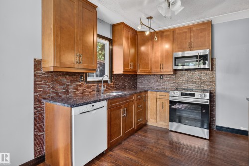 Kitchen featuring wood cabinetry, stainless steel appliances, a tile backsplash, dark countertops, and wood-finish flooring - 13351 109A Avenue Nw, Edmonton, AB 
