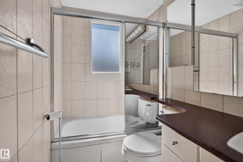 Floor-to-ceiling tiled walls, enclosed bathtub with sliding glass doors, horizontal transom window, extended vanity with integrated sink, and overhead multi-bulb lighting fixture - 13351 109A Avenue Nw, Edmonton, AB 