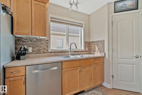 Wood-finish cabinetry with stainless steel hardware, stone-finish countertops, stainless steel dishwasher, mosaic tile backsplash, and a double basin sink with gooseneck faucet - 591 Byrne Crescent Sw, Edmonton, AB 