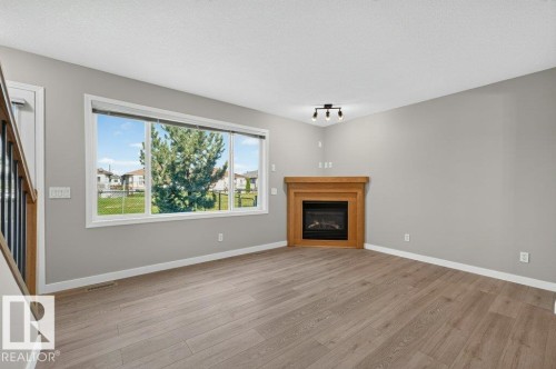 Spacious room featuring wood-finish flooring, a contemporary fireplace with a wood mantle, and a large multi-pane window - #26 2816 34 Av Nw, Edmonton, AB 