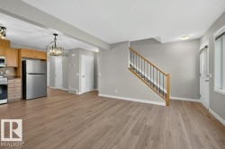 Open-concept living area with wood-finish flooring, a kitchen featuring stainless steel appliances and wood cabinetry, a staircase with wood handrails, and modern lighting fixtures - 
