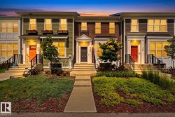 Townhome exterior featuring varied siding materials including brick and horizontal plank siding, multiple entryways with individual staircases and black railings, ground-level landscaping with mulch and low-lying shrubs, and upper-level window planters - 