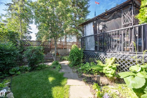 Backyard featuring a paved pathway, manicured lawn, established garden beds, and a raised wooden deck with black railings and lattice skirting - 11007 108 Avenue, Westlock, AB 