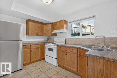 Kitchen featuring wood-finish cabinetry, stainless steel refrigerator, white range, and a dual-basin sink with a gooseneck faucet - 14527 32 Street Nw, Edmonton, AB 