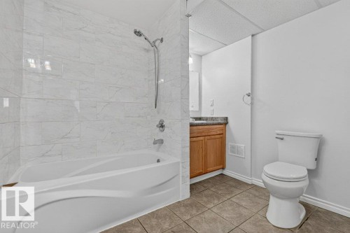 White tiled shower surround, built-in bathtub, wood-finish vanity with counter, ceramic tile flooring, and a white toilet - 14527 32 Street Nw, Edmonton, AB 