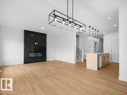 Wood-finish flooring throughout, modern linear chandelier, floor-to-ceiling black stone-finish fireplace, kitchen island with light wood-finish cabinetry, and contemporary pendant lighting - 912 17 Ave Nw, Edmonton, AB 