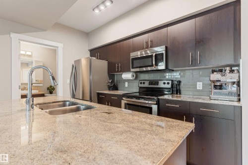 Kitchen featuring an island with double stainless steel sink and gooseneck faucet, light-toned granite countertops, dark wood-finish cabinetry, and stainless steel appliances including a refrigerator, range, and microwave - #2205 10226 104 St Nw, Edmonton, AB - Indoor Photo Showing Kitchen With Double Sink With Upgraded Kitchen
