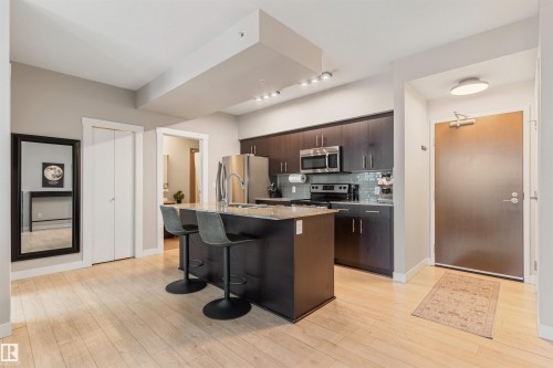 Open-concept kitchen featuring an island with seating, stainless steel appliances, dark wood-finish cabinetry, a tiled backsplash, and light wood-finish flooring - #2205 10226 104 St Nw, Edmonton, AB - Indoor Photo Showing Kitchen With Upgraded Kitchen