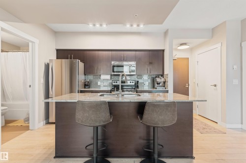 Kitchen island featuring a light-toned stone countertop, integrated sink with a stainless steel faucet, and dark wood-finish cabinetry - #2205 10226 104 St Nw, Edmonton, AB - Indoor Photo Showing Kitchen With Upgraded Kitchen