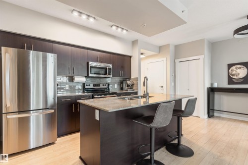 Modern kitchen featuring a central island, stainless steel appliances, dark wood-finish cabinetry, a tile backsplash, and light wood-finish flooring - #2205 10226 104 St Nw, Edmonton, AB - Indoor Photo Showing Kitchen With Double Sink With Upgraded Kitchen