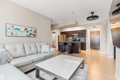 Open-concept living area featuring light wood-finish flooring, a contemporary kitchen with dark wood cabinetry, a stainless steel appliance suite, and a breakfast bar with pendant lighting - #2205 10226 104 St Nw, Edmonton, AB - Indoor Photo Showing Living Room