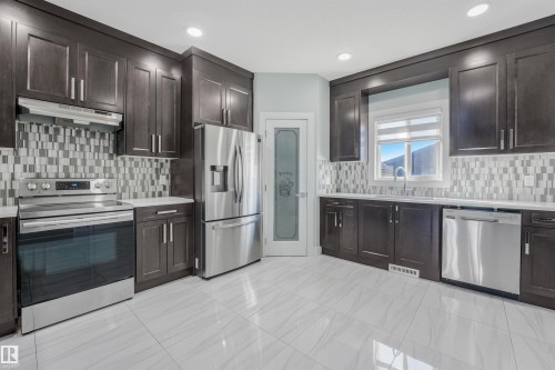 Kitchen featuring dark wood-finish cabinetry, stainless steel appliances, a mosaic tile backsplash, white countertops, and light-toned tile flooring - 6453 177A Avenue Nw, Edmonton, AB 