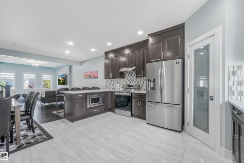 Contemporary kitchen featuring dark wood cabinetry, stainless steel appliances, a tiled backsplash, a kitchen island with built-in microwave, and large format floor tiling - 6453 177A Avenue Nw, Edmonton, AB 