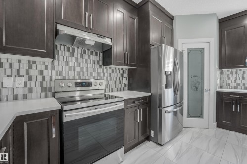 Kitchen featuring dark wood-finish cabinetry, stainless steel appliances, a mosaic tile backsplash, light-toned countertops, and a pantry with a frosted glass door - 6453 177A Avenue Nw, Edmonton, AB 