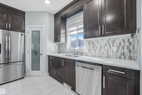 Contemporary kitchen featuring dark wood cabinetry, stainless steel appliances, a mosaic tile backsplash, and light-toned countertops - 6453 177A Avenue Nw, Edmonton, AB 