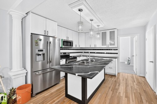 Kitchen featuring wood-finish flooring, stainless steel appliances, white cabinetry, a black and white striped tile backsplash, and a central island with a sink and dark countertop - Edmonton, AB 