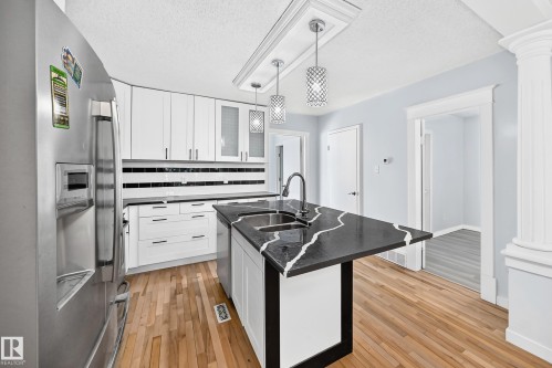 Kitchen featuring white cabinetry, a black and white patterned island with an undermount sink, stainless steel refrigerator, wood-finish flooring, and pendant lighting - Edmonton, AB 
