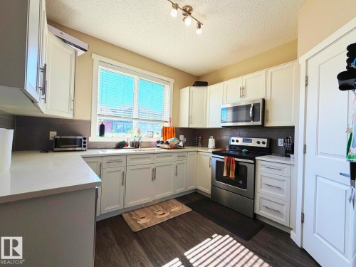 Kitchen featuring white cabinetry, light-toned countertops, stainless steel appliances, a dark tile backsplash, and wood-finish flooring - 5169 Chappelle Rd Southwest, Edmonton, AB 