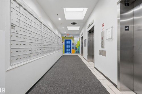 Modern building corridor featuring wall-mounted mailboxes with individual compartments, polished stainless steel elevator doors, recessed ceiling lighting, neutral wall finishes, and gray tile flooring throughout - #1313 10024 Jasper Av Nw, Edmonton, AB 