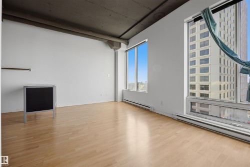 Expansive room featuring light wood-finish flooring, high ceilings with exposed concrete, and large windows offering urban surroundings - #1313 10024 Jasper Av Nw, Edmonton, AB 