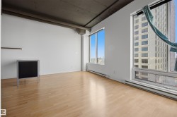 Expansive room featuring light wood-finish flooring, high ceilings with exposed concrete, and large windows offering urban surroundings - 